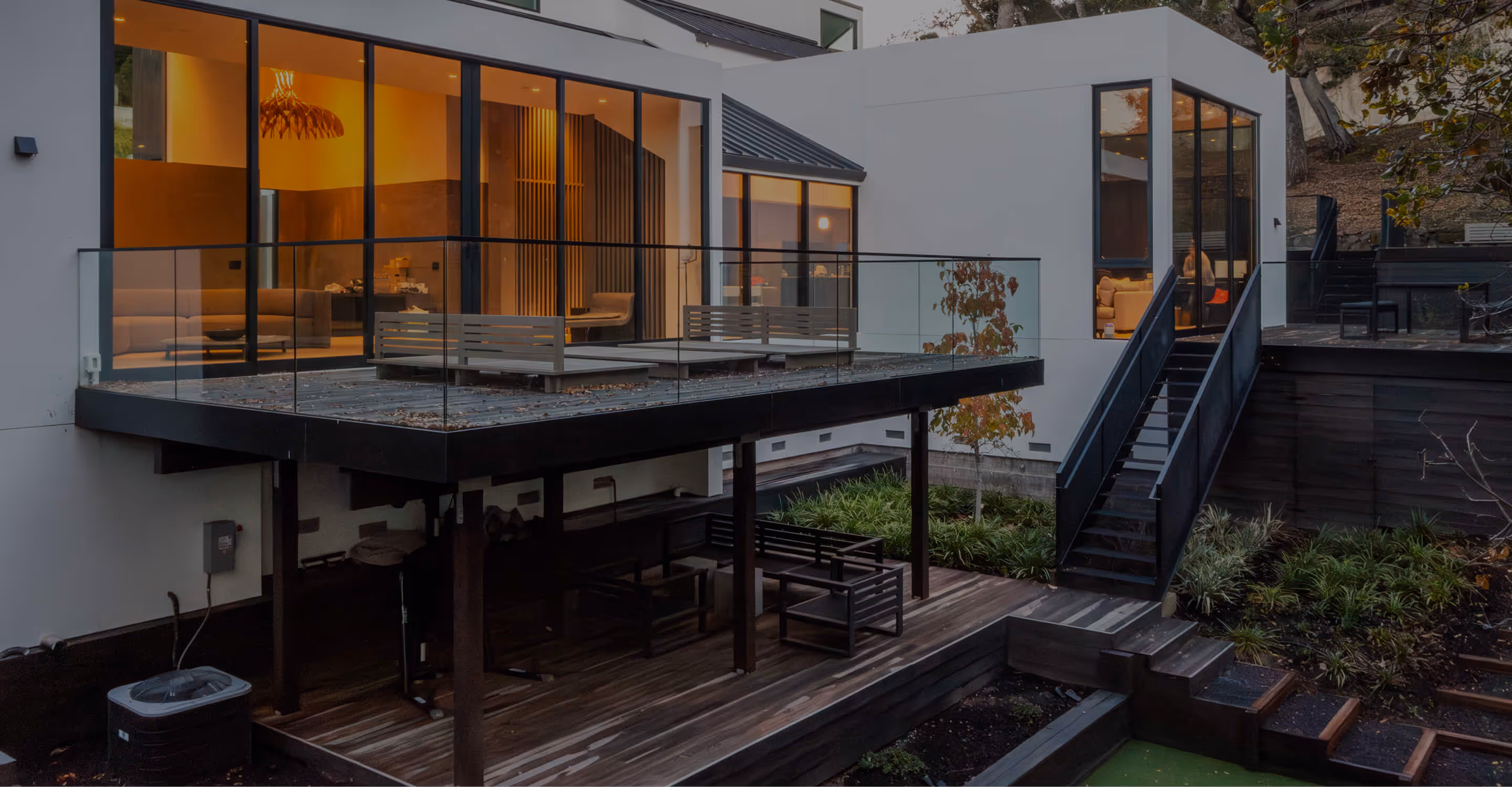 Modern house exterior with a glass-railed upper deck and wooden lower patio with outdoor seating, surrounded by greenery.