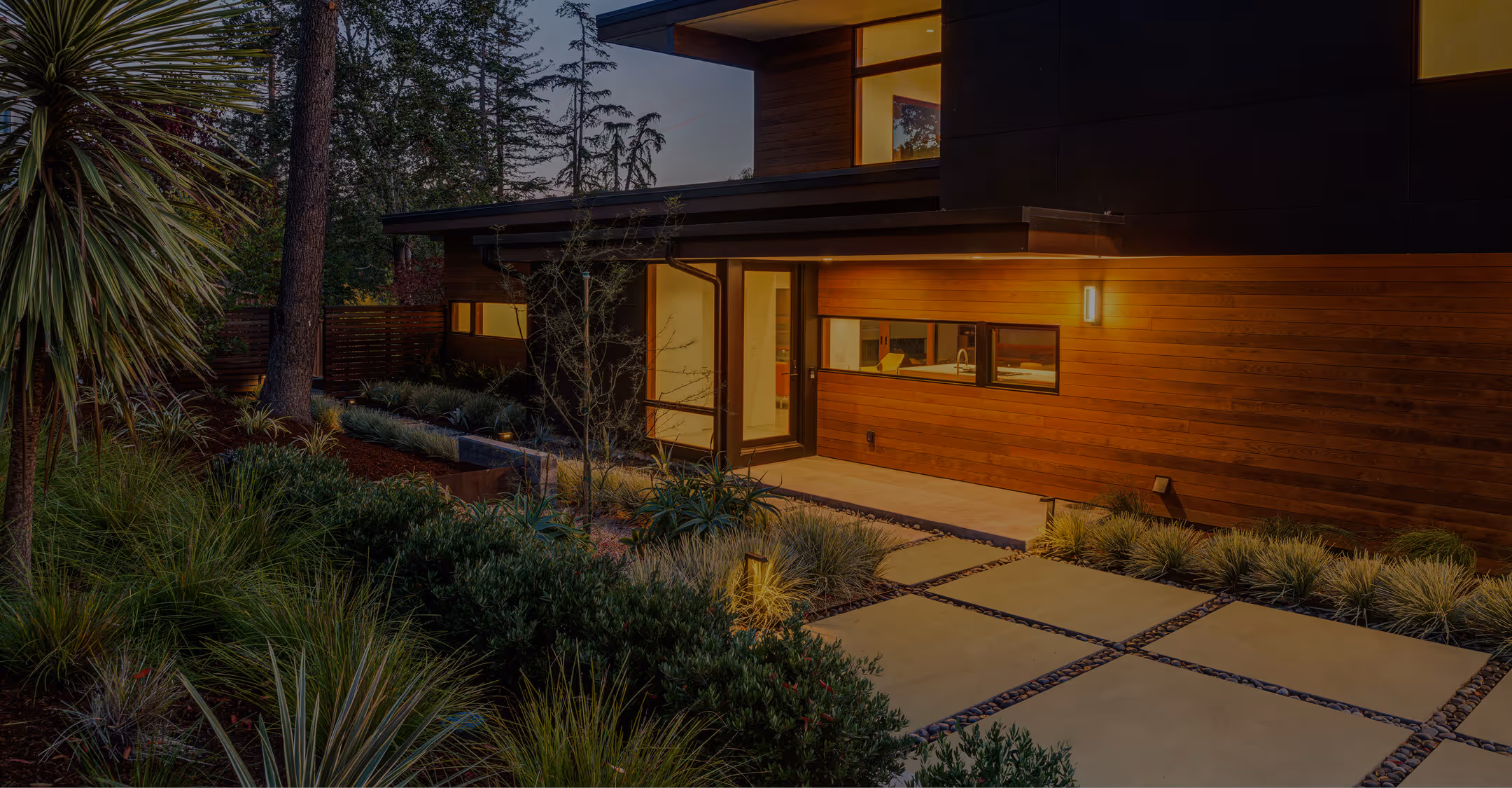 Modern house exterior with wooden paneling, large windows, lit entrance, and landscaped garden with concrete tile pathway.
