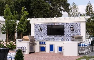 Outdoor kitchen setup with a grill, blue patterned tile backsplash, wall-mounted TV, and seating with bar stools.