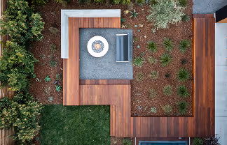 Aerial view of a backyard patio with wooden decking, a built-in bench, a fire pit, and surrounding greenery.