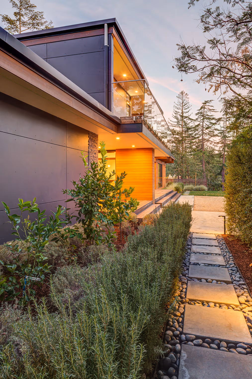 Modern house exterior with a stone pathway, landscaped greenery, and a second-floor balcony with glass railing illuminated at dusk.