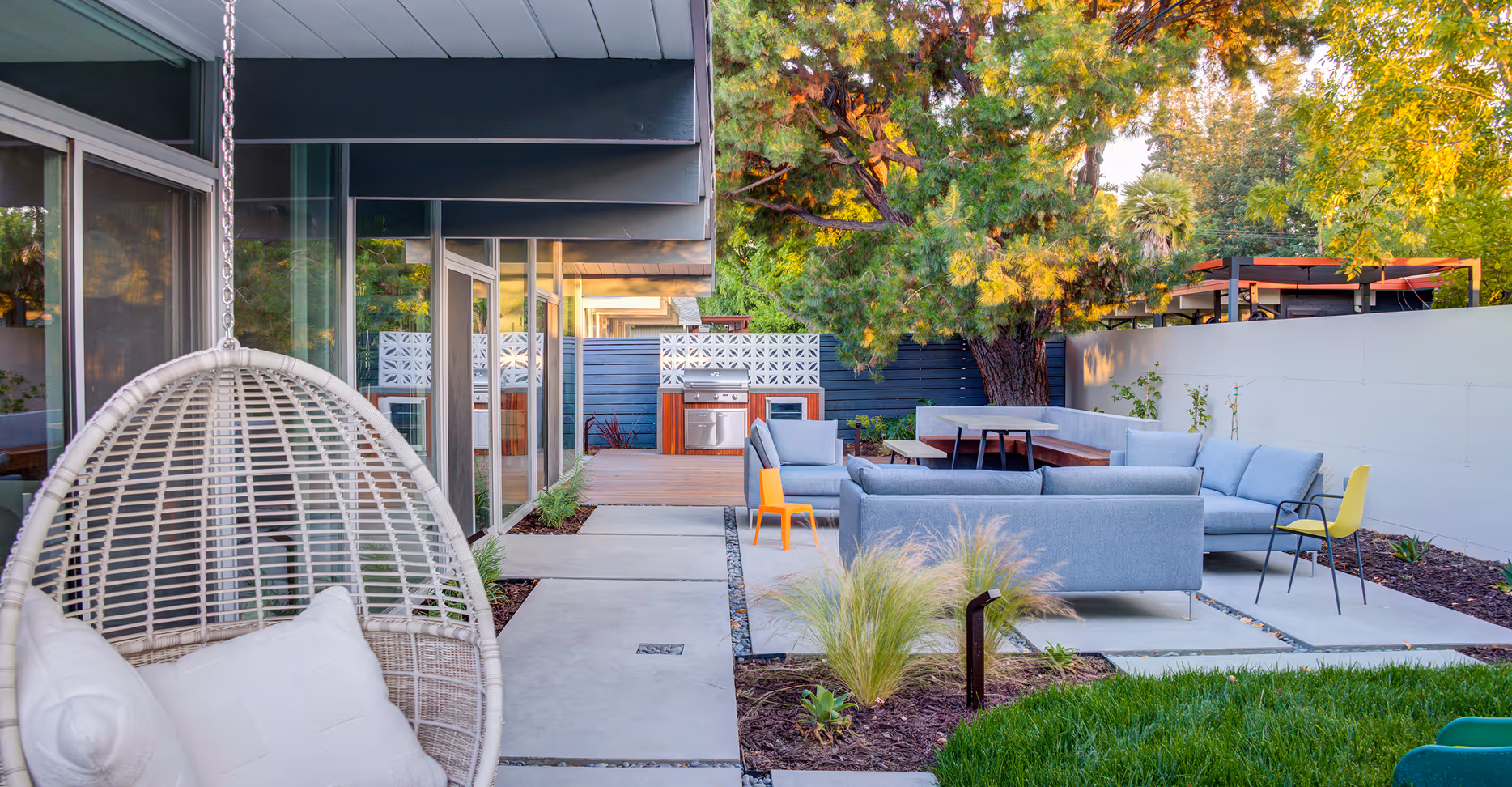 Modern backyard patio with outdoor kitchen, gray sofas, hanging wicker chair, and lush greenery.