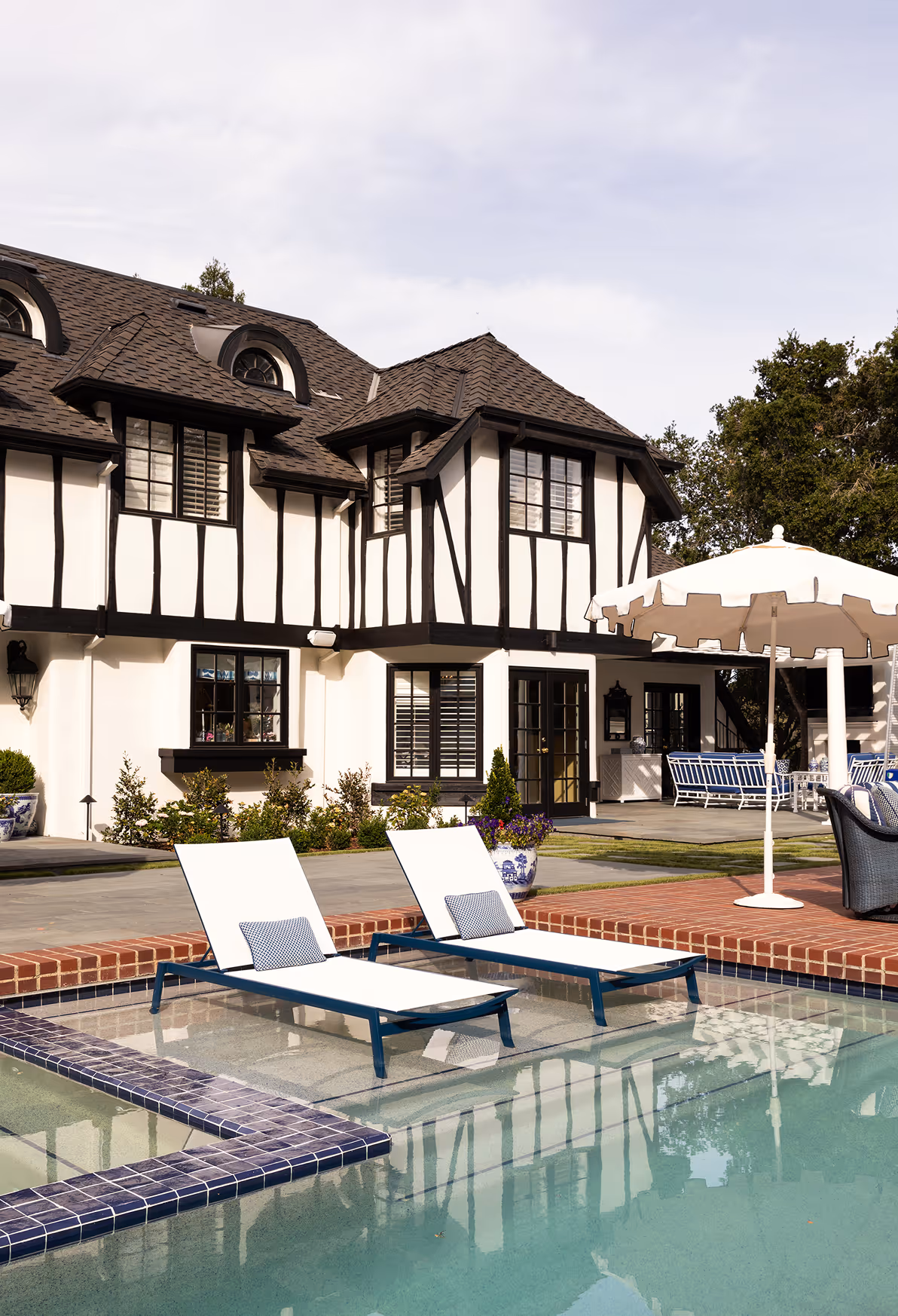 Two white lounge chairs with blue cushions partially submerged in a pool next to a black and white Tudor-style house patio with an umbrella and outdoor seating.