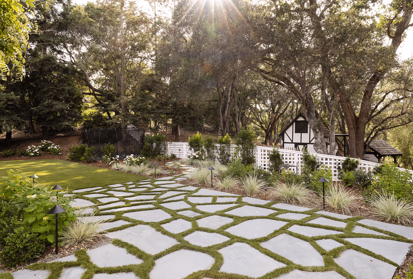 Sunlit garden with large irregular stone path, green lawn, white lattice fence, and a small Tudor-style house surrounded by trees.