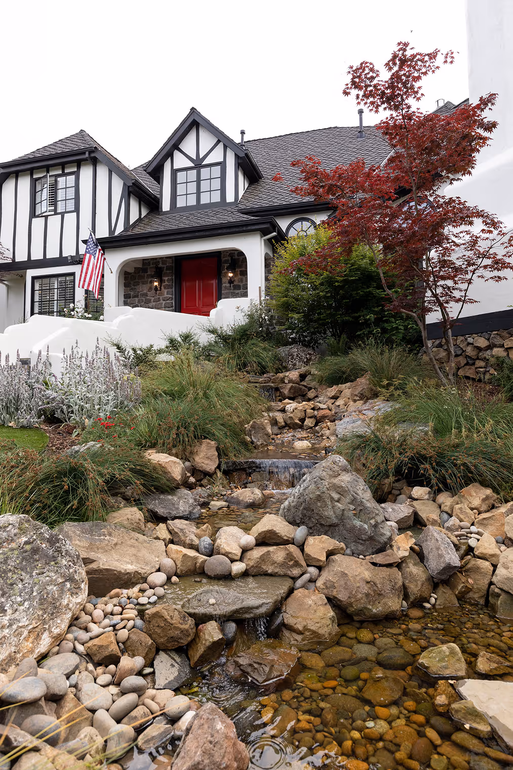 Tudor-style house with a red door, an American flag, and a landscaped yard featuring a small rock-lined stream and various plants.