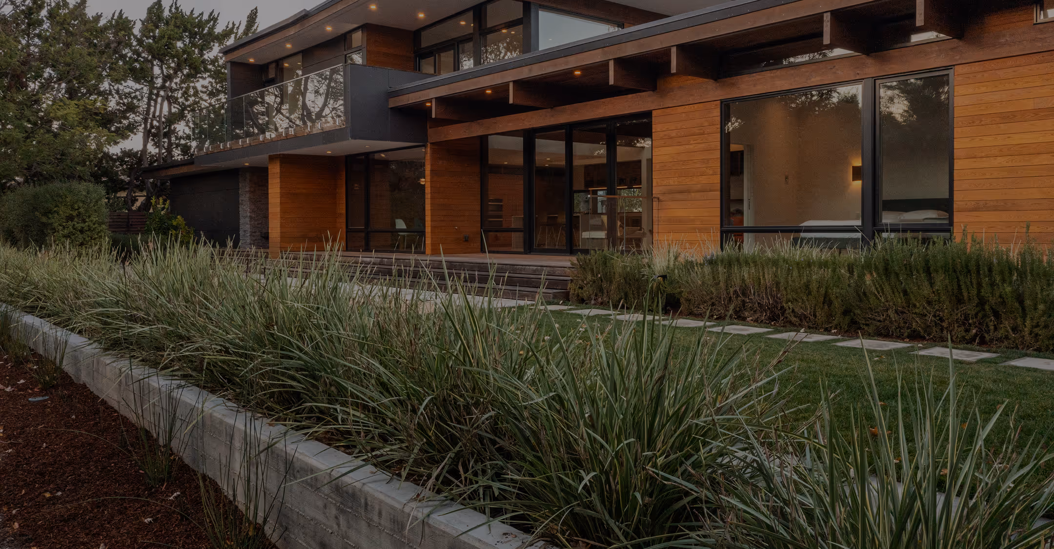 Modern wooden house with large glass windows and an elevated balcony surrounded by landscaping.
