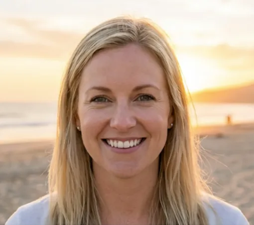 Smiling blonde woman standing on a beach at sunset.