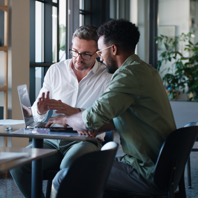 Two men sitting at a table, engaged in a discussion while looking at a laptop screen in a modern office setting.