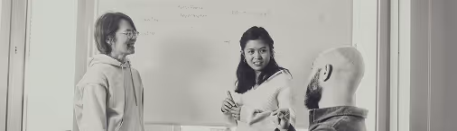Three people engaged in a discussion in front of a whiteboard, two standing and one sitting.