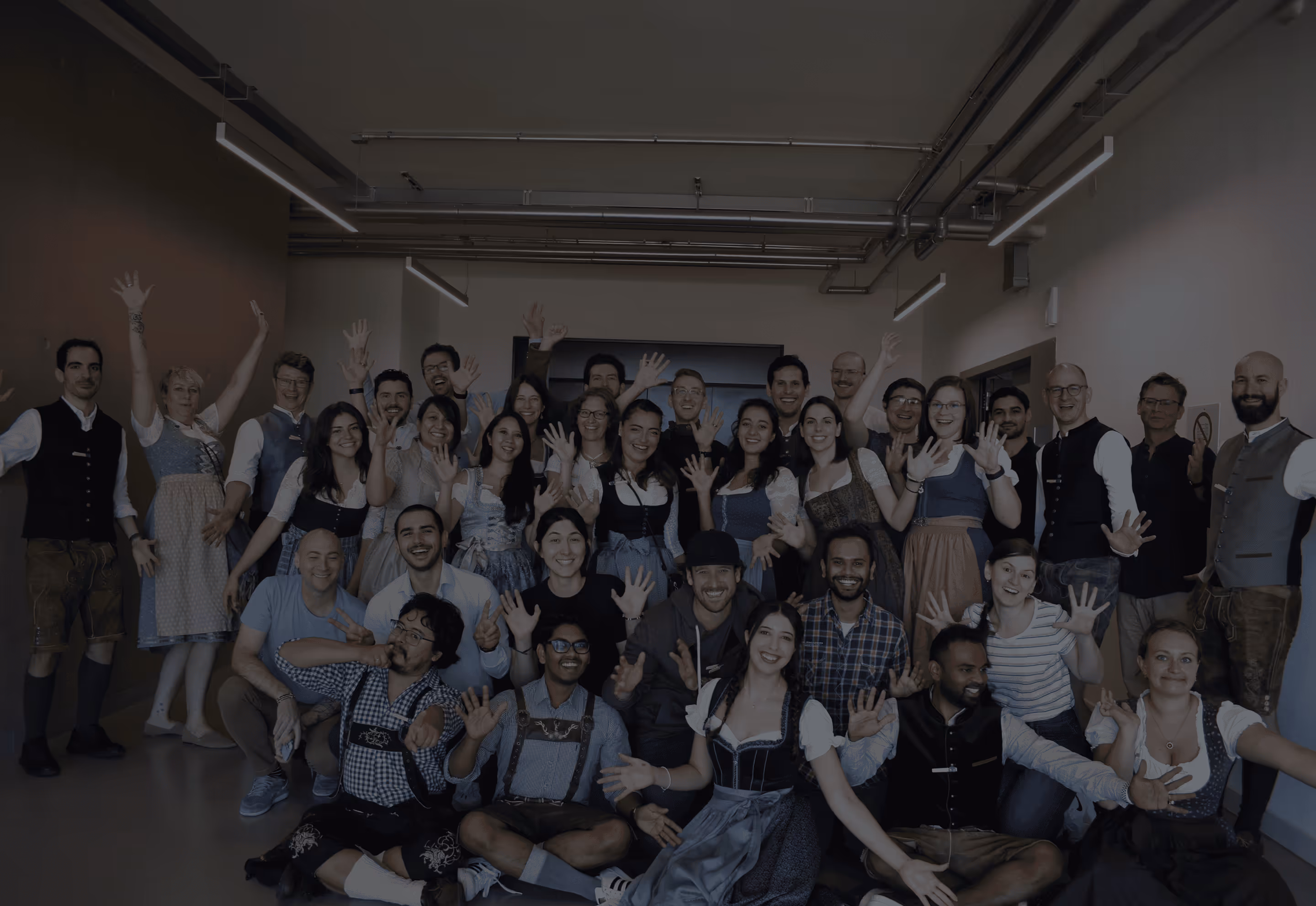Large group of people smiling and waving at the camera in an indoor setting, many wearing traditional Bavarian clothing.
