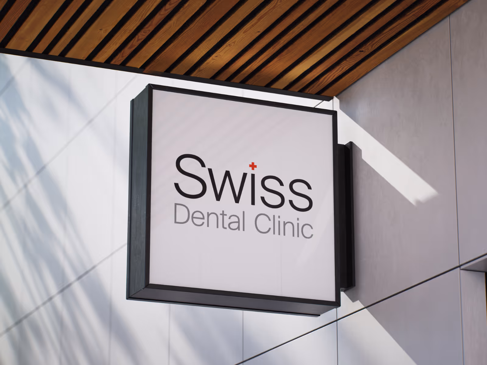 Square signboard mounted on a wall displaying 'Swiss Dental Clinic' with a small red cross above the letter 'i' in Swiss.