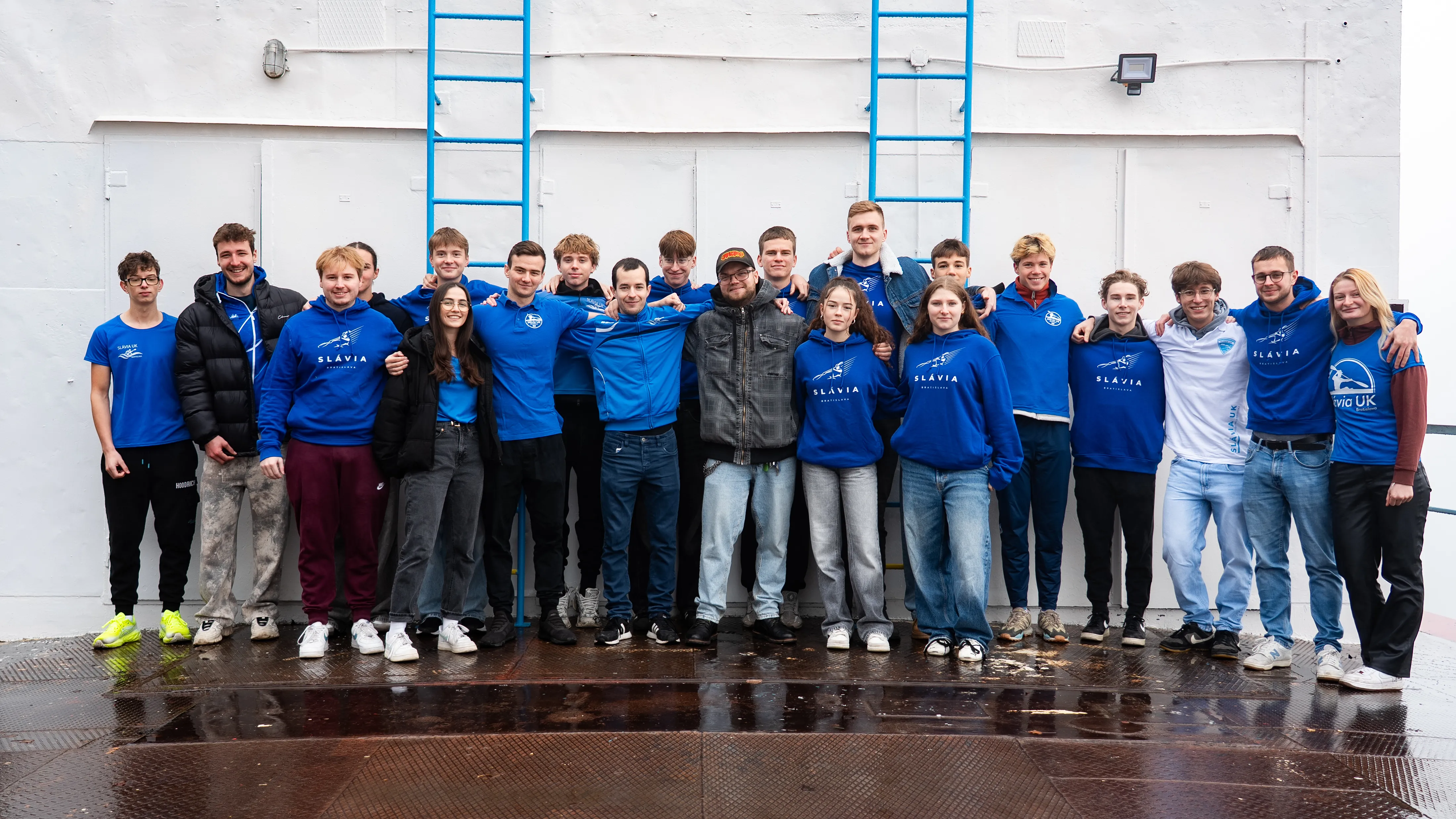 Group of 20 young adults standing close together against a white wall with blue ladder rungs, most wearing blue Slávia team apparel.