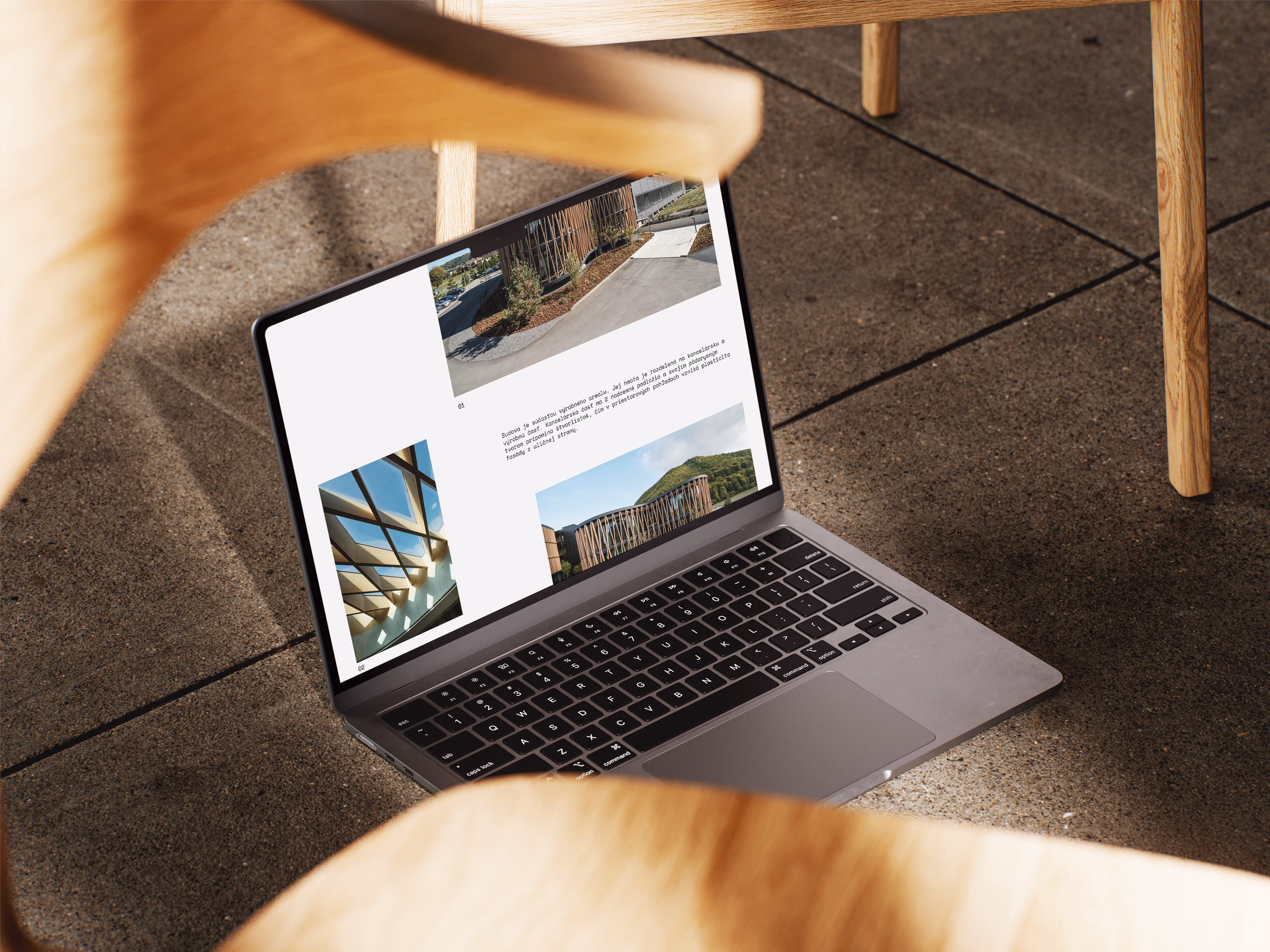 Open laptop on a concrete floor showing a webpage with images of architectural outdoor structures, surrounded by wooden chair legs.