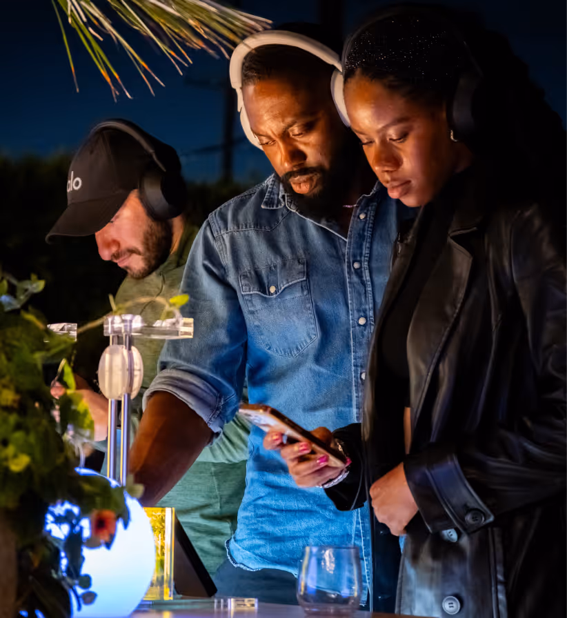 Three people wearing headphones closely looking at a smartphone under soft outdoor lighting at night.