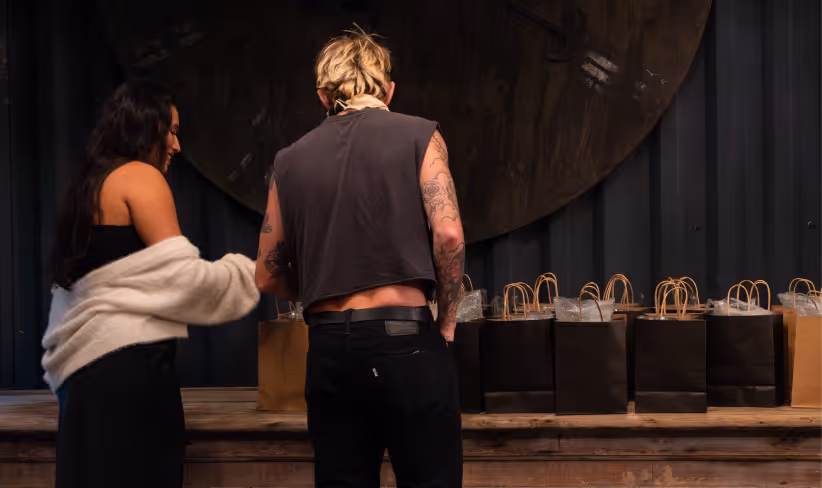 Two people standing at a wooden table with multiple black and brown gift bags arranged on it.
