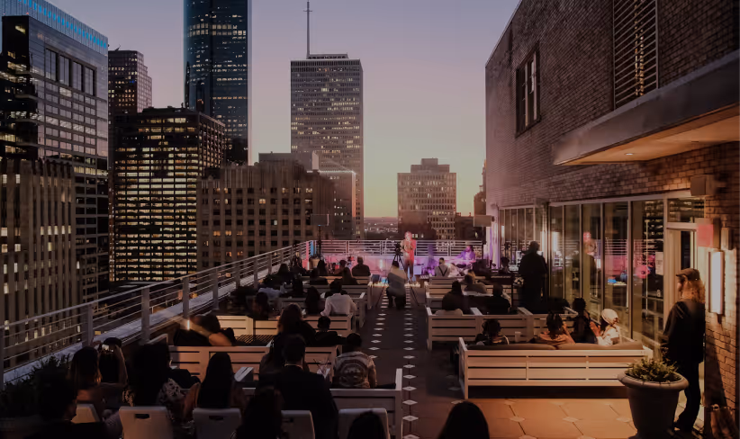 People seated on benches at a rooftop event during sunset with city skyscrapers in the background.
