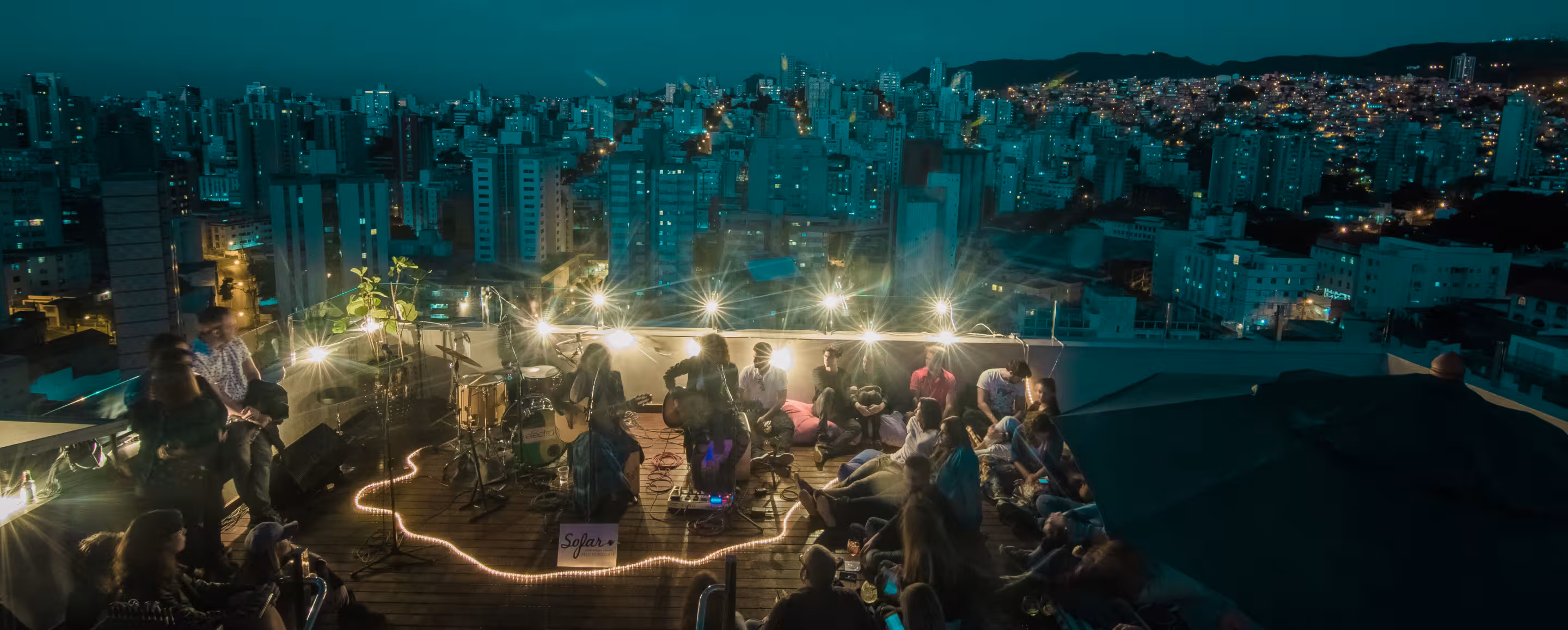 Nighttime rooftop concert with two acoustic guitarists and an audience against a city skyline backdrop.