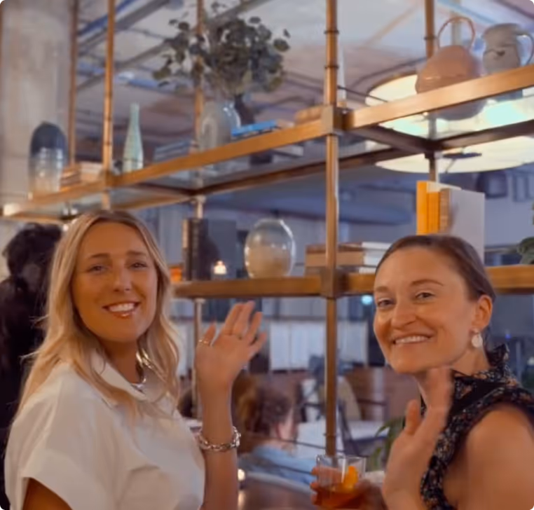 Two women smiling and waving while holding drinks in a cozy indoor setting with shelves and plants behind them.