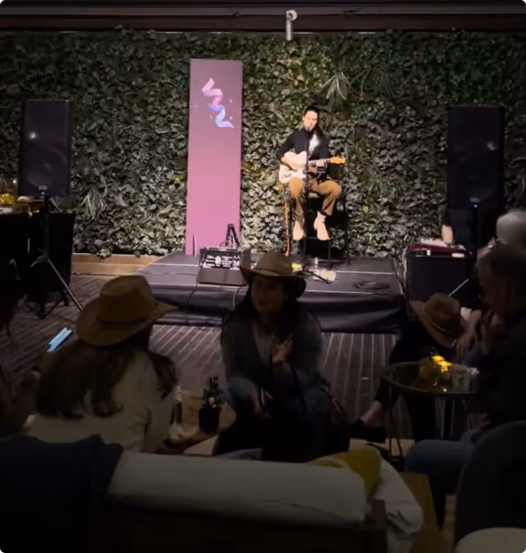 Musician playing guitar and singing on small stage with leafy backdrop in dimly lit venue with seated audience.