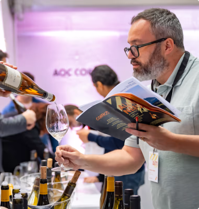 Man with glasses holding a booklet and receiving white wine poured into a glass at a tasting event.