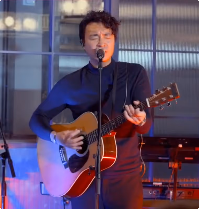Man in black shirt singing and playing an acoustic guitar on a dimly lit stage.