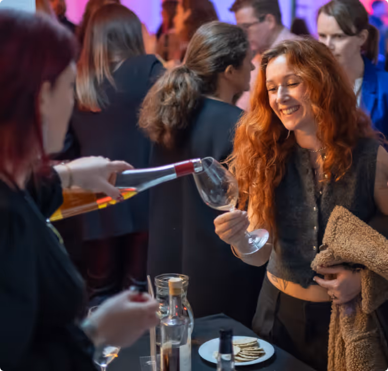 Smiling red-haired woman holding a wine glass while another person pours wine at a social event.