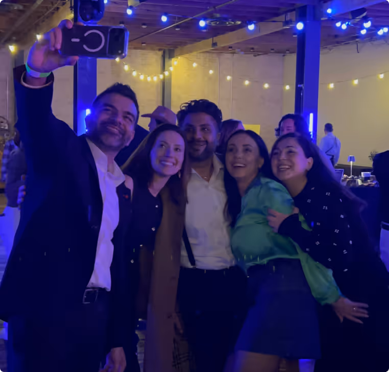 Group of five people smiling and posing for a selfie at an indoor event with string lights overhead.