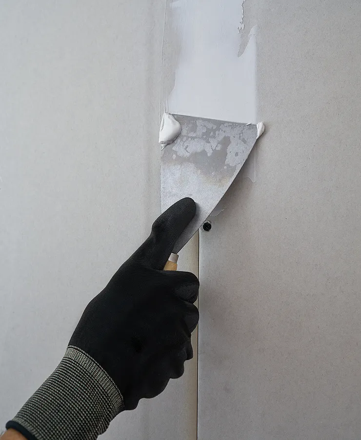 Hand wearing a black glove applying drywall joint compound with a putty knife on a drywall seam.