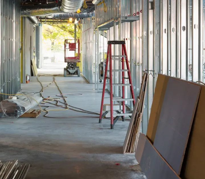 Interior view of a building under construction with metal framing, a red ladder, scattered tools, and construction materials on a concrete floor.