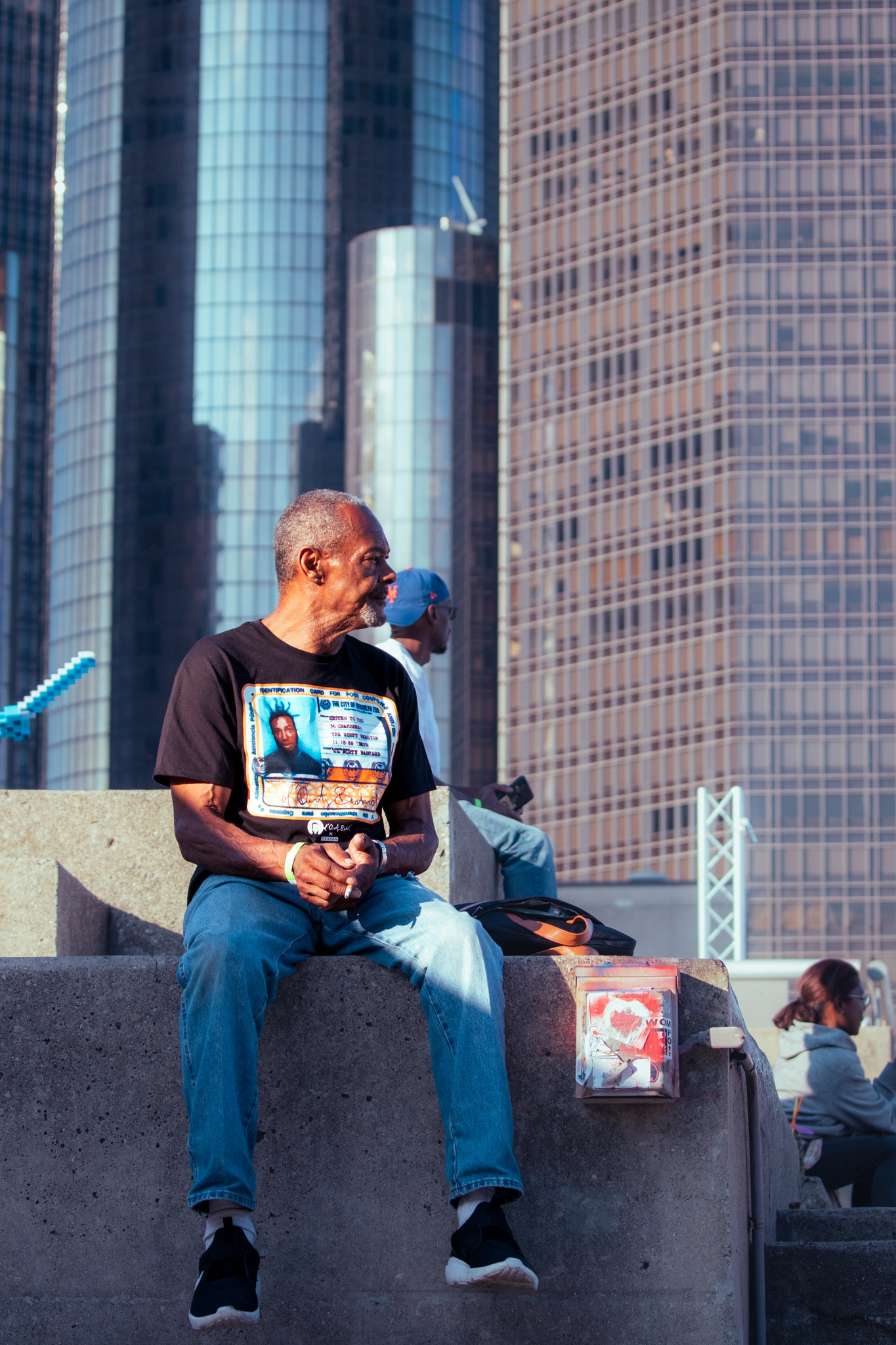 Older man in black graphic t-shirt and jeans sitting on a concrete ledge with tall glass buildings in the background at sunset.