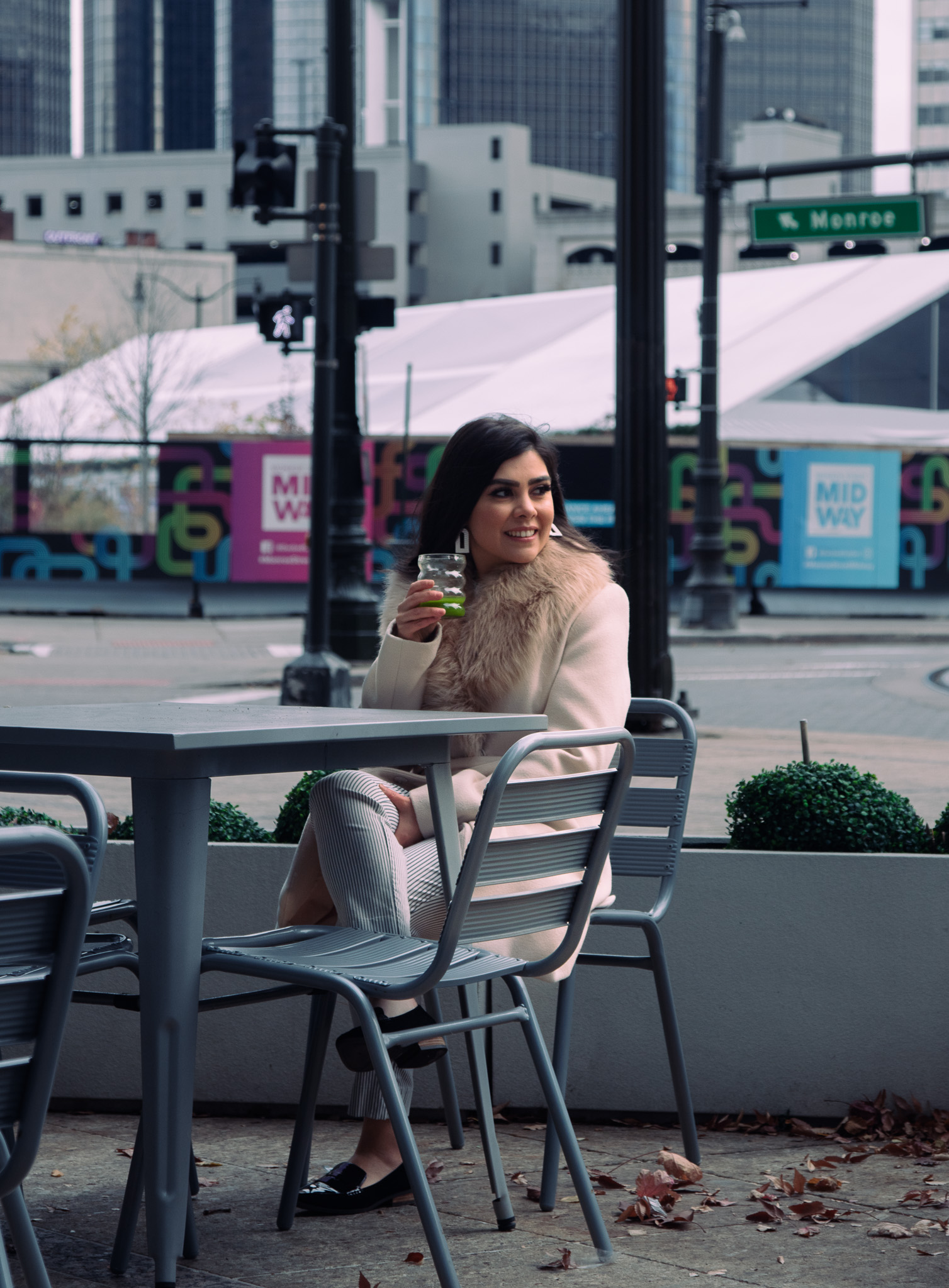 Woman wearing a beige coat with fur collar sitting outdoors at a gray metal table holding a glass with green drink, urban background with street signs and buildings.