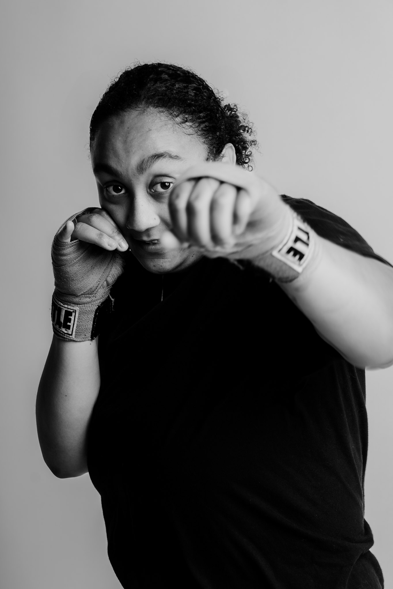 Person in black shirt with wrapped hands throwing a punch toward the camera against a plain background.