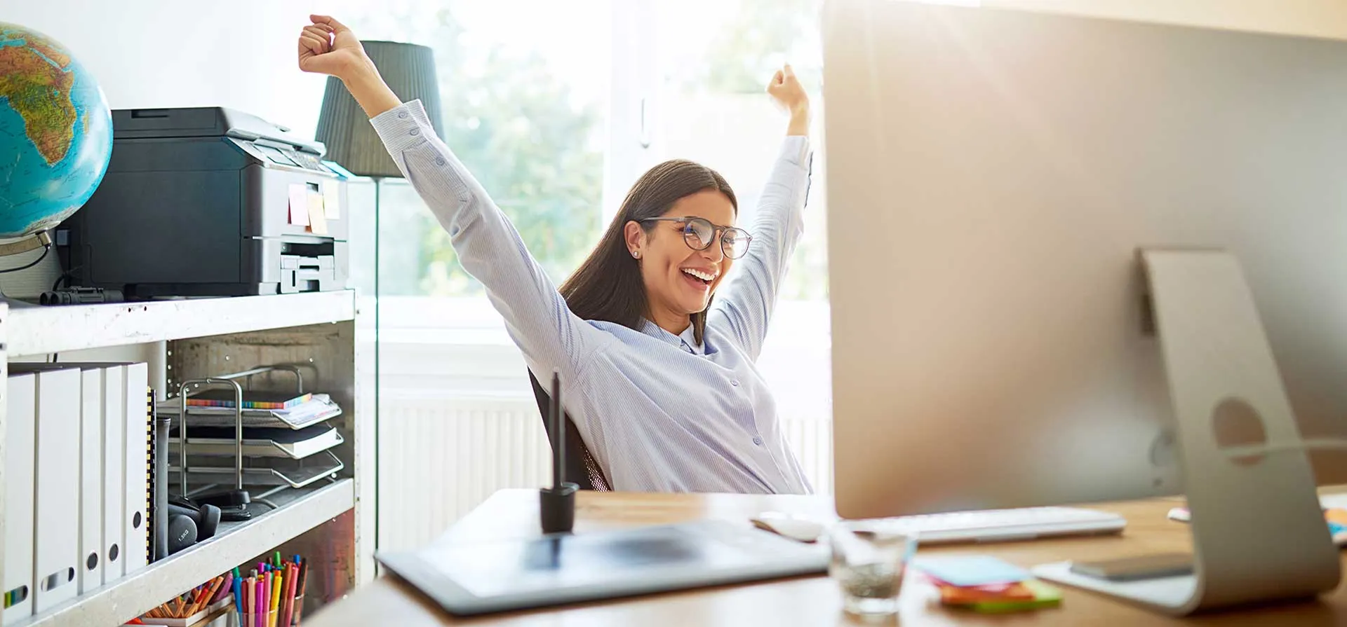 Smiling woman stretching with raised arms while sitting at a desk with a computer in a bright office.