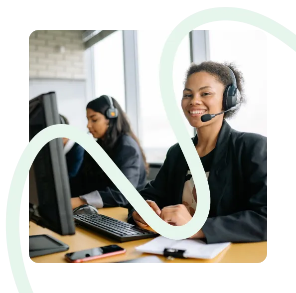 Two women wearing headsets working at computers in an office, one smiling at the camera.