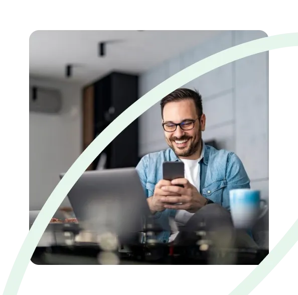 Smiling man with glasses sitting at a table using a smartphone with a laptop and coffee cup nearby.