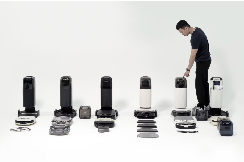 Man in black shirt demonstrating multiple robotic vacuum cleaners and their dustbins and accessories arranged in a row on the floor against a white background.
