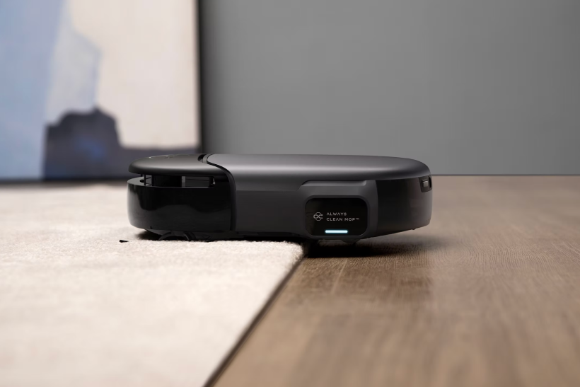 Black robot vacuum cleaner positioned on the edge of a carpet and wooden floor, displaying 'Always Clean Mop' on its side screen.