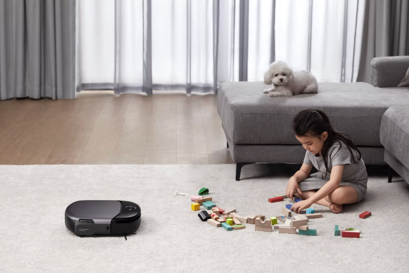 A young girl sitting on a carpeted floor playing with colorful wooden blocks while a small white dog lies on a gray ottoman in a modern living room with sheer curtains.