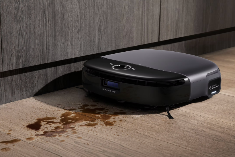Black robotic vacuum cleaner on wooden floor near dark wall, cleaning a liquid spill.