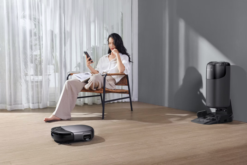 Woman sitting on a chair reading a book and holding a smartphone, with a robot vacuum cleaner operating on a wooden floor and a charging station against a gray wall.