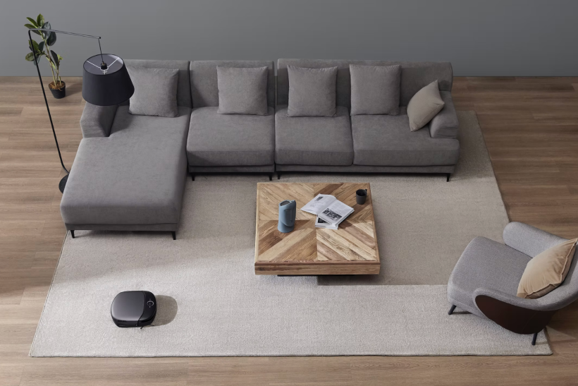 Modern living room with a gray sectional sofa, a wooden coffee table holding books and a pitcher, a gray armchair with a beige pillow, a floor lamp, a potted plant, and a robotic vacuum cleaner on a beige rug over wooden floor.
