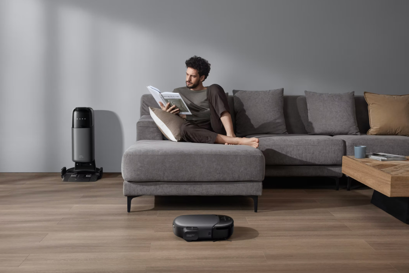 Man sitting on a gray sectional sofa reading a book with a robotic vacuum cleaner on the wooden floor and its charging station nearby.