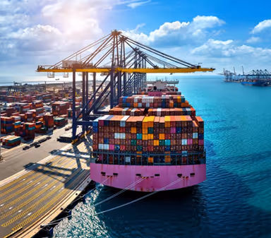 Large container ship docked at a port with stacked colorful shipping containers and cranes under a partly cloudy sky.