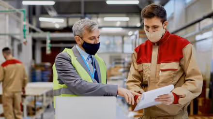Two factory workers wearing masks and discussing documents in an industrial setting.
