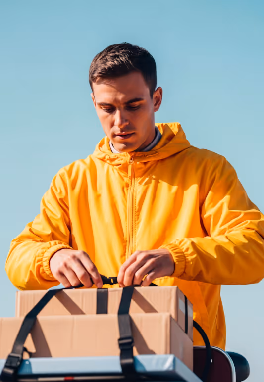 Man in yellow jacket securing cardboard boxes with black straps outdoors under clear blue sky.