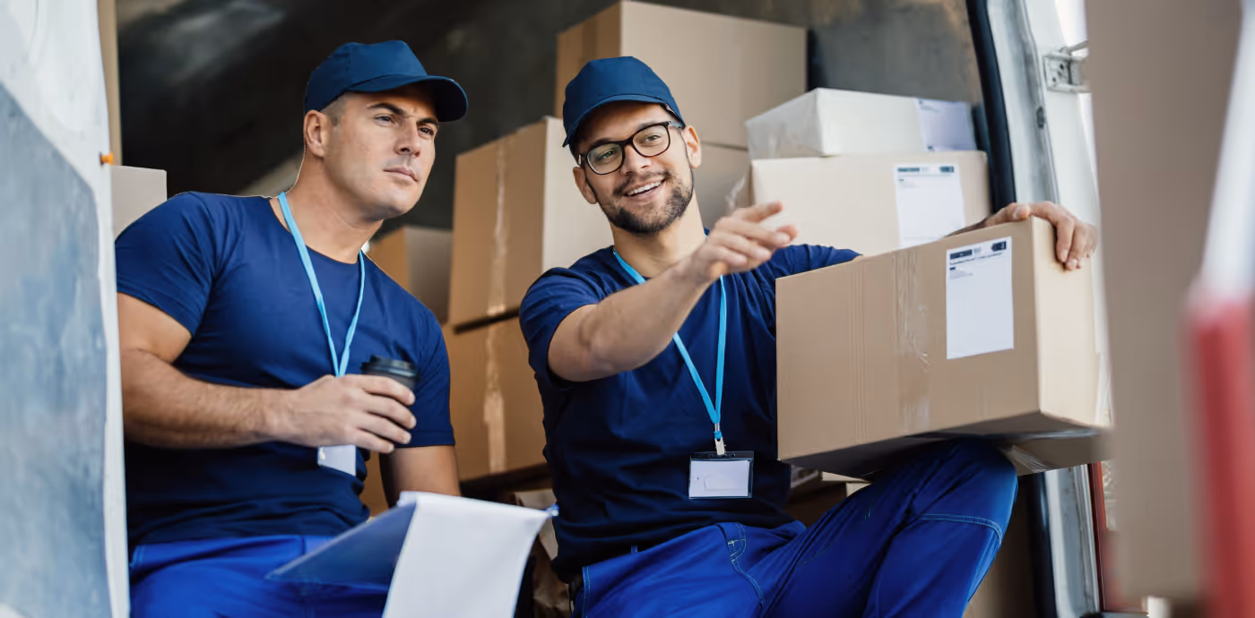 Two delivery workers in blue uniforms sitting in a van with cardboard boxes, one holding a coffee cup and clipboard, the other holding and pointing with a package.