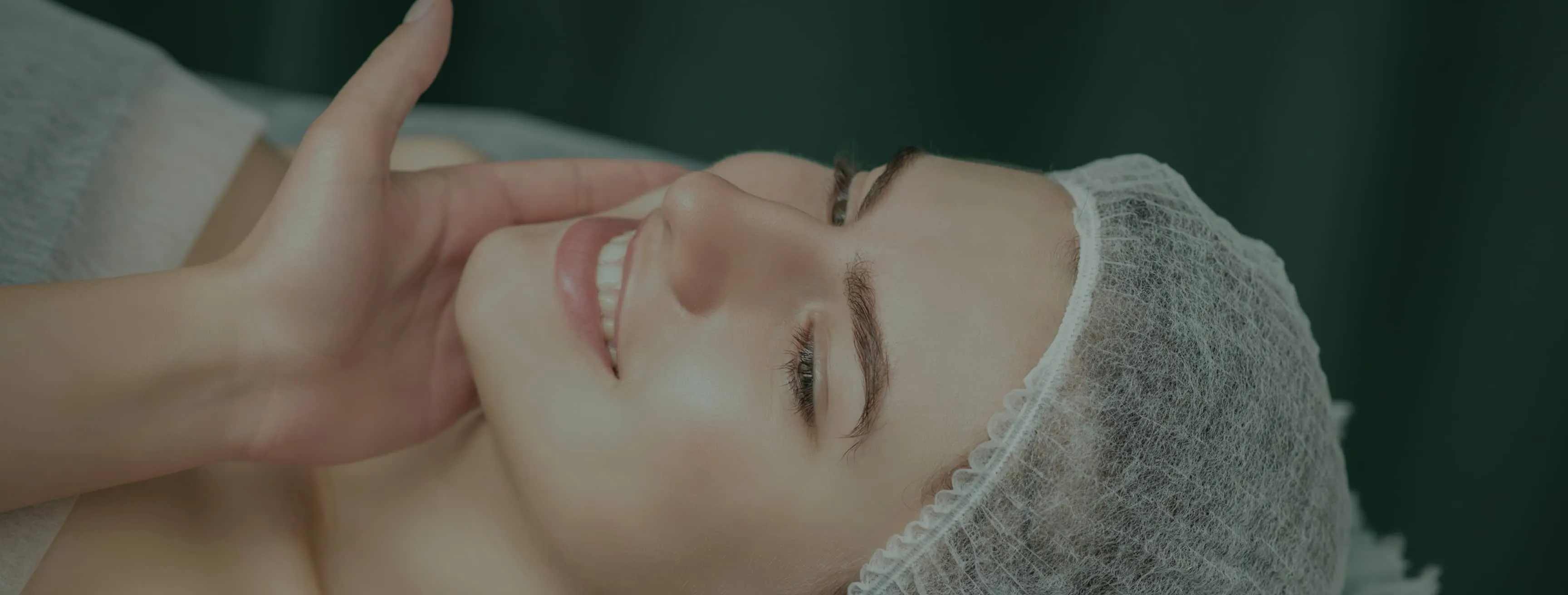 Close-up of a smiling woman with smooth skin wearing a hairnet, resting her face on her hand against a dark background.