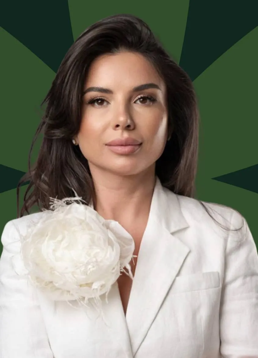 Woman with long dark hair wearing a white blazer adorned with a large white floral accessory on the shoulder, against a dark green background with radiating segments.