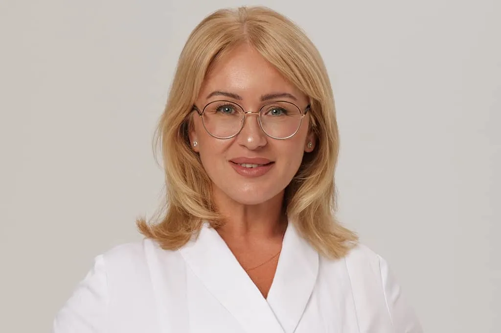 Smiling middle-aged woman with blonde hair and glasses wearing a white lab coat.