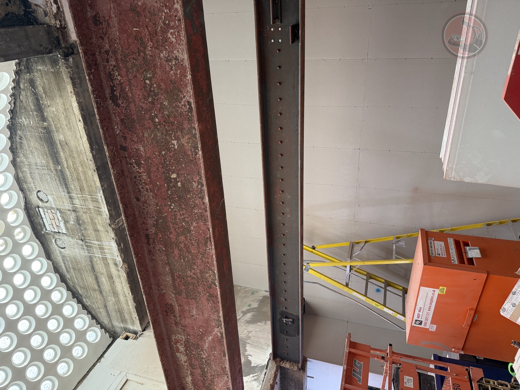 Indoor construction scene showing two steel beams, one old rusted and one painted, with a yellow ladder and orange equipment in front of a white paneled wall.